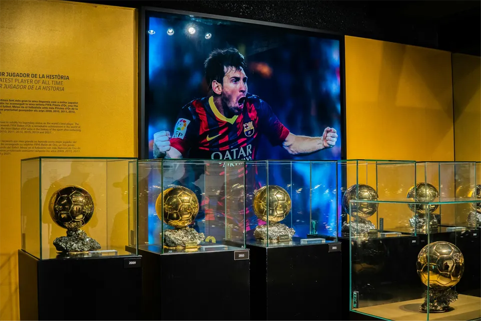 Lionel Messi's Ballon d'Or trophies in glass cases beneath a large photograph of him celebrating in a Barcelona shirt
