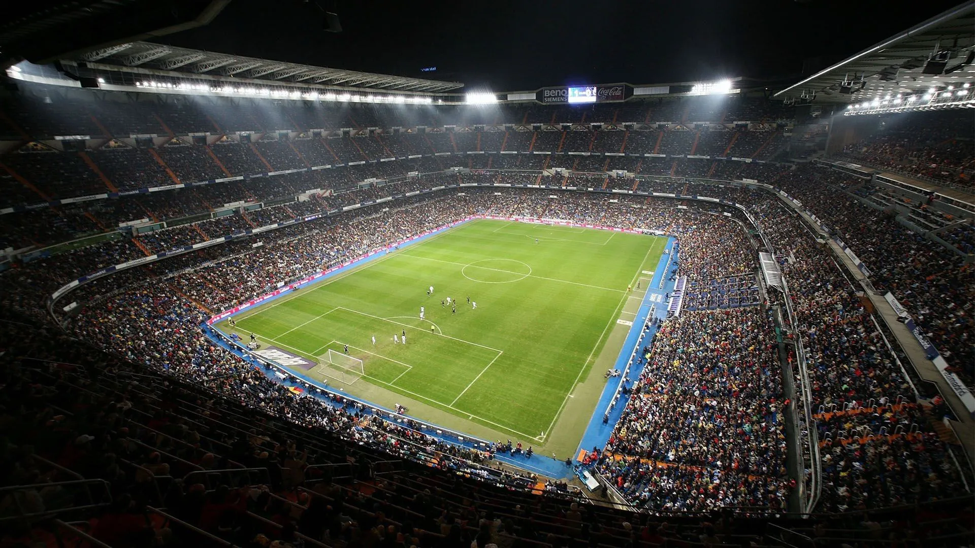 A packed football stadium under floodlights, seen from high in the stand